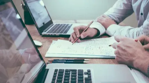 Nella foto si intravedono due persone sedute ad un tavolo, davanti ai propri notebook, che discutono di questioni di lavoro prendendo note su un foglio.